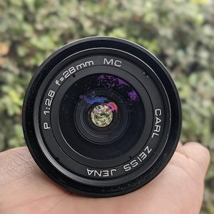 May include: A black Carl Zeiss Jena camera lens with the text "P 1:2.8 f=28mm MC" and "CARL ZEISS JENA" printed on the lens. The lens is held in a hand, with a blurred green background.