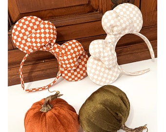 May include: Two pairs of checkered fabric mouse ears headbands in orange and white and beige and white. Three velvet pumpkins in orange, green, and brown.