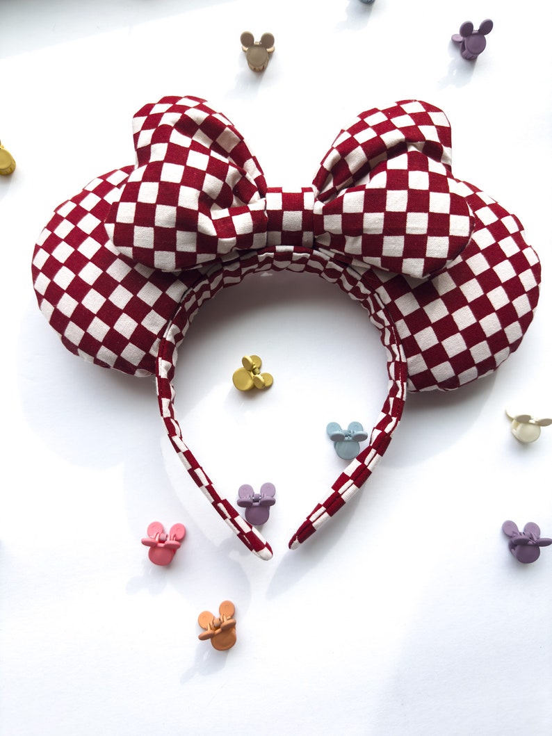 May include: Red and white checkered fabric headband with a large bow. The headband is shaped like Mickey Mouse ears.