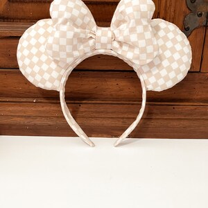 May include: A pair of beige and white checkered mouse ears with a large bow. The ears are attached to a headband.