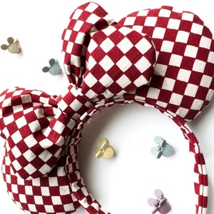 May include: Red and white checkered fabric headband with a large bow. The headband is designed to look like Mickey Mouse ears.