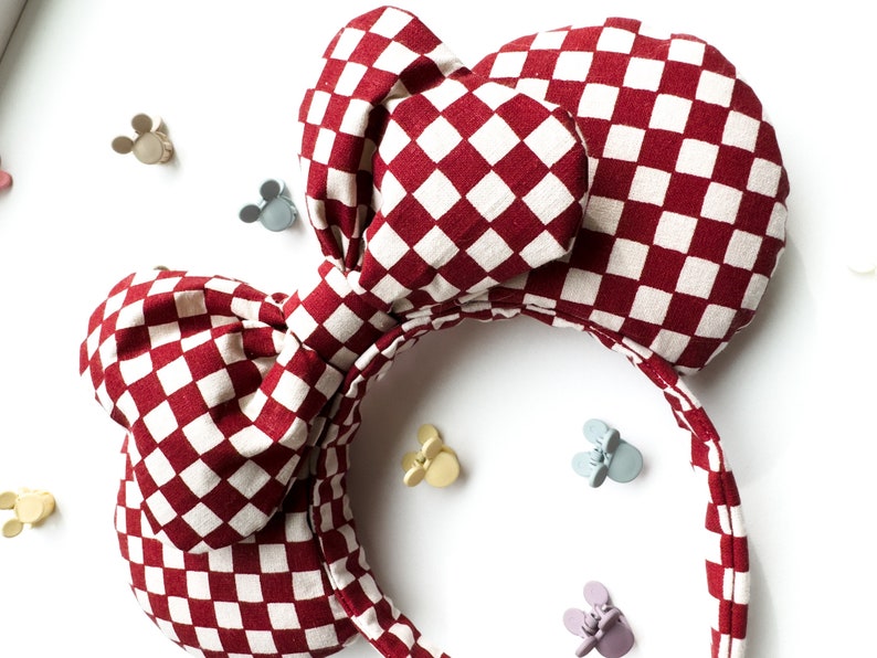 May include: Red and white checkered fabric headband with a large bow. The headband is designed to look like Mickey Mouse ears.