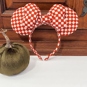 May include: Red and white checkered fabric mouse ears headband with a large bow. The headband is on a wooden surface.