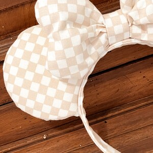 May include: A pair of beige and white checkered mouse ears with a large bow. The headband is beige and white checkered.