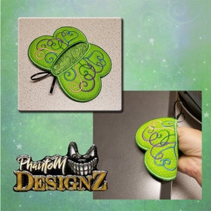 Könnte beinhalten: Ein grüner herzförmiger Topflappen mit einem schwarzen Band und einem Wirbelmuster. Der Topflappen ist aus Stoff und hat ein weißes Punkte-Muster. Der Topflappen ist auf einer grauen Oberfläche abgebildet.