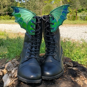 Puede incluir: Botas de cuero negro con alas de dragón verde iridiscente atadas a los cordones. Las alas son de tela y tienen una textura escamosa.