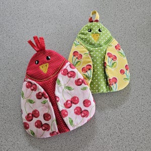 Peut inclure: Deux maniques en forme d'oiseau. L'une est rouge à pois blancs et ailes à motifs de cerises. L'autre est verte à pois blancs et ailes à motifs de cerises jaunes. Les deux ont des becs jaunes et des boucles de suspension.