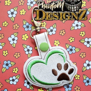 May include: White glitter heart-shaped keychain with a green embroidered outline and a brown paw print in the center. The keychain has a green button and a silver metal ring.