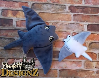 May include: Two plush stingray toys. One is a dark blue color, and the other is white with gray wings. Both have embroidered eyes and smiling faces. The logo "Phantom Designz" is at the bottom.