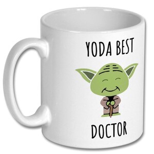 May include: White ceramic coffee mug featuring a green Yoda character holding a walking stick. The text "Yoda Best Doctor" is printed on the mug.