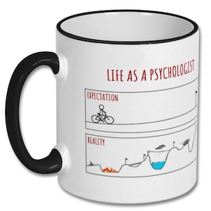 May include: White ceramic mug with a black rim and handle. The mug features a humorous illustration comparing the expectation and reality of being a psychologist. The text reads "Life as a Psychologist".