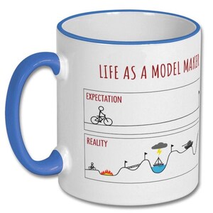 May include: White ceramic mug with a blue handle and rim. The mug features the text "LIFE AS A MODEL MAKER" in red, along with a humorous illustration comparing expectations to reality, including a cyclist and a tank.