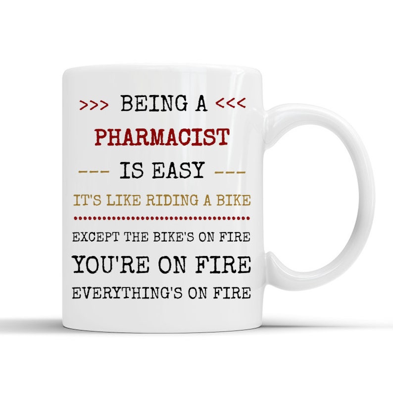 May include: White ceramic coffee mug with a humorous saying about being a pharmacist. The text reads: >>> BEING A <<< PHARMACIST --- IS EASY --- IT'S LIKE RIDING A BIKE EXCEPT THE BIKE'S ON FIRE YOU'RE ON FIRE EVERYTHING'S ON FIRE