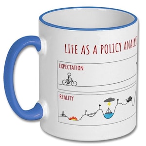 May include: White ceramic mug with a blue handle and rim. The mug features the text "LIFE AS A POLICY ANALYST" in red, along with a humorous graphic comparing expectations to reality, including a cyclist and a boat.