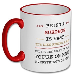 May include: White ceramic coffee mug with a red rim and handle. The mug has a humorous quote printed on it: ">>> BEING A <<< SURGEON IS EASY IT'S LIKE RIDING A BIKE EXCEPT THE BIKE'S ON FIRE YOU'RE ON FIRE EVERYTHING'S ON FIRE"
