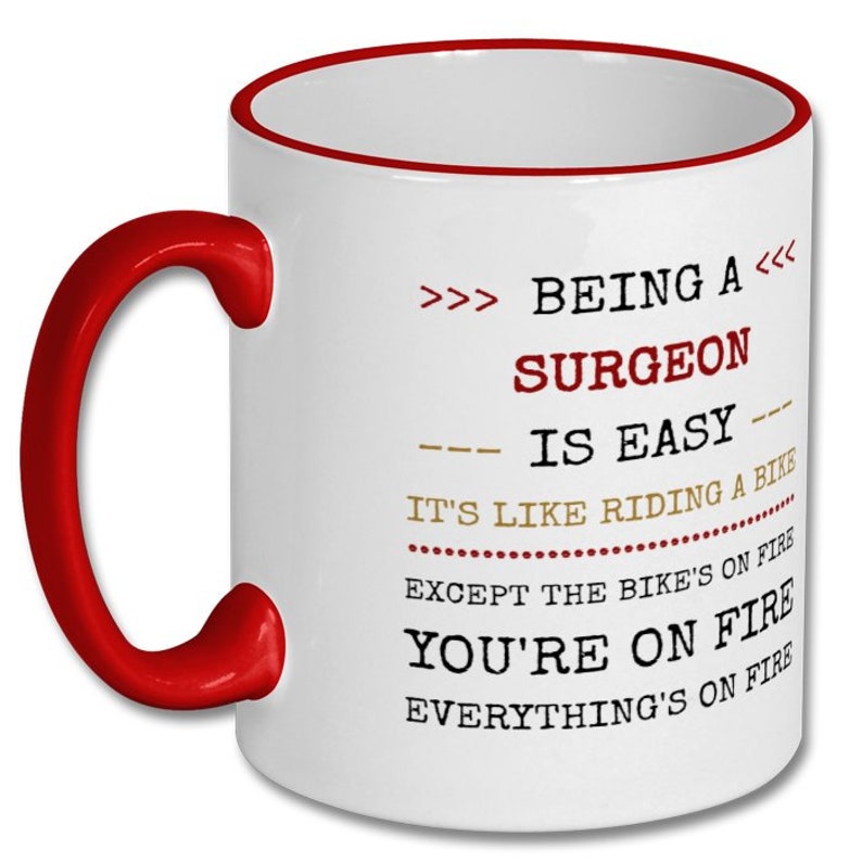 May include: White ceramic coffee mug with a red rim and handle. The mug has a humorous quote printed on it: ">>> BEING A <<< SURGEON IS EASY IT'S LIKE RIDING A BIKE EXCEPT THE BIKE'S ON FIRE YOU'RE ON FIRE EVERYTHING'S ON FIRE"