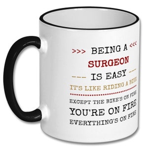 May include: White coffee mug with a black rim and handle. The mug has a humorous quote about being a surgeon: ">>> BEING A <<< SURGEON IS EASY --- IT'S LIKE RIDING A BIKE EXCEPT THE BIKE'S ON FIRE YOU'RE ON FIRE EVERYTHING'S ON FIRE"