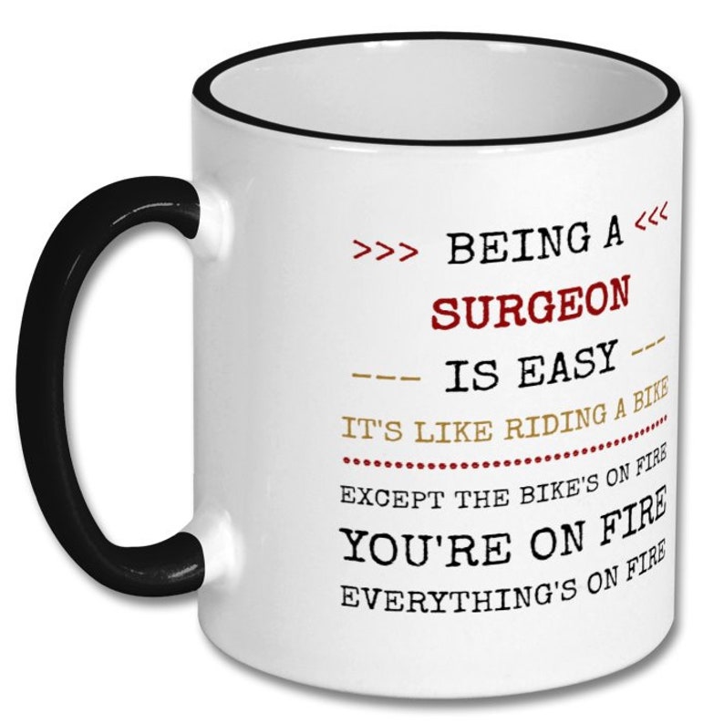 May include: White coffee mug with a black rim and handle. The mug has a humorous quote about being a surgeon: ">>> BEING A <<< SURGEON IS EASY --- IT'S LIKE RIDING A BIKE EXCEPT THE BIKE'S ON FIRE YOU'RE ON FIRE EVERYTHING'S ON FIRE"