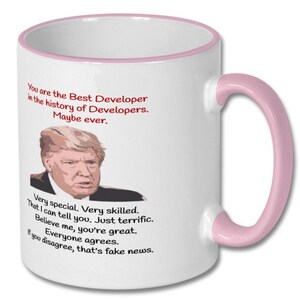 May include: White coffee mug with a pink rim and handle. The mug features a cartoon image of Donald Trump and the text "You are the Best Developer in the history of Developers. Maybe ever. Very special. Very skilled. That I can tell you. Just terrific. Believe me, you're great. Everyone agrees. If you disagree, that's fake news."