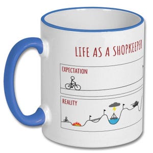 May include: White ceramic mug with a blue handle and rim. The mug features the text "LIFE AS A SHOPKEEPER" in red, along with a humorous illustration comparing expectations to reality, including a cyclist and a tank.