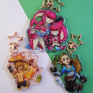 May include: Three colorful keychains featuring characters from the Legend of Zelda video game series. The first keychain features a pink and blue character with a fish-like tail. The second keychain features a character with a sword and a fire background. The third keychain features a character with a blue dress and a frog.