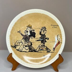 May include: A decorative plate with a cream-colored rim and a tan center featuring a cartoon illustration of people walking. The plate has the text "Promenade à Paris". The plate is displayed on a wooden stand.