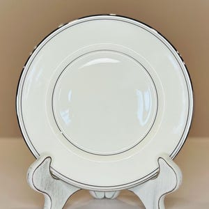May include: A white ceramic plate with a black and silver trim, displayed on a distressed white wooden stand. The plate has a simple, elegant design, suitable for formal dining. The stand has a decorative, curved shape.