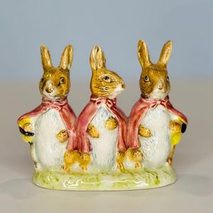 May include: A ceramic figurine featuring three anthropomorphic rabbits. Each rabbit wears a pink cape and holds a yellow object. The rabbits stand on a green base, with a light blue background. The figurine is likely a decorative item.