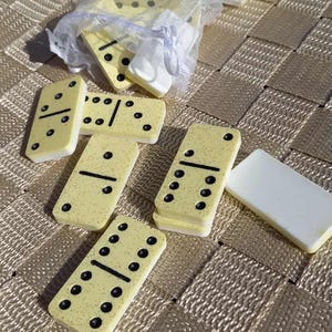 May include: A collection of yellow and white dominoes with black dots. Some dominoes are scattered, while others are in a sheer bag tied with a ribbon. The dominoes are on a textured, gold-colored surface.