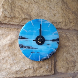 May include: A blue and white painted clock with black hands, featuring a seascape with rocks and water. The clock is mounted on a stone wall.