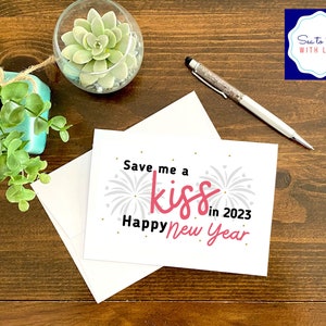 May include: A white card with a pink and black design that says "Save me a kiss in 2023 Happy New Year". The card is on a wooden table with a small potted plant and a pen.