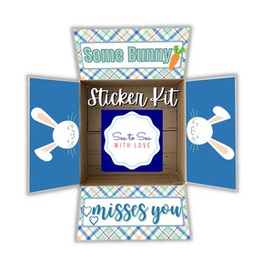 May include: A blue and white plaid box with the text "Some Bunny" and a carrot on the top flap. The box is open and the inside is blue with a white bunny face. The bottom flap has the text "misses you" with two hearts. The box also has a sticker with the text "Sea to Sea with Love".