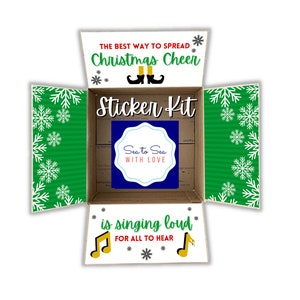 May include: An open cardboard box with a green and white snowflake pattern on the sides. The box is labeled "The Best Way to Spread Christmas Cheer" and "Sticker Kit". The box also includes a blue and white sticker with the text "Sea to Sea With Love". The bottom of the box has the text "is singing loud for all to hear" with a musical note.