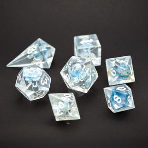 Könnte beinhalten: Ein Set aus sieben transparenten Harzwürfeln mit blauen Blumeneinschlüssen, angeordnet auf einer schwarzen Oberfläche. Die Würfel haben verschiedene Formen, darunter eine Pyramide, ein Würfel und ein 20-seitiger Würfel, mit weißen Zahlen.