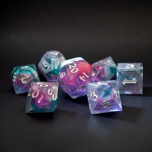 Op de afbeelding: Een verzameling van zeven polyhedrale dobbelstenen met een galaxy-achtig ontwerp. De dobbelstenen hebben verschillende vormen, waaronder D4, D6, D8, D10, D12 en D20. De kleuren zijn teal, paars, roze en wit, met zilveren nummering.
