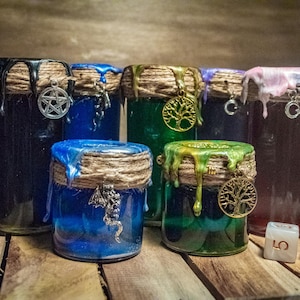 Könnte beinhalten: Sechs Glasgefäße mit verschiedenen farbigen Flüssigkeiten und Wachstropfen. Jedes Gefäß hat ein Band aus Schnur und Jute um die Oberseite und einen Anhänger, der an dem Band hängt. Die Anhänger umfassen einen Pentagramm, einen Lebensbaum und einen Halbmond. Die Gefäße stehen auf einer Holzoberfläche mit drei weißen Würfeln.