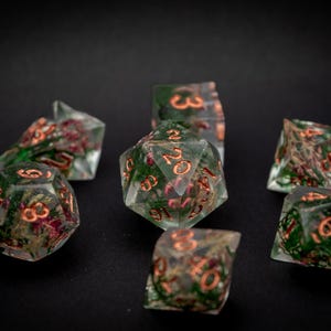 Könnte beinhalten: Sieben polyedrische Würfel mit transparentem Harz, grünen und roten Einschlüssen. Die Zahlen sind kupferfarben. Geeignet für Tabletop-Rollenspiele.