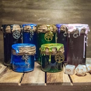 Könnte beinhalten: Sechs Glasgefäße mit verschiedenen farbigen Wachsen und Charms. Die Gefäße sind mit Schnur verziert und haben eine rustikale, magische Ästhetik. Die Gefäße sind auf einer Holzoberfläche mit weißen Würfeln angeordnet.