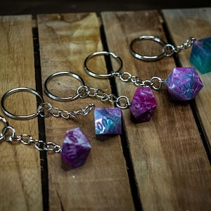 May include: Five colorful resin dice keychains with silver key rings and chains. The dice are in shades of pink, purple, and blue.