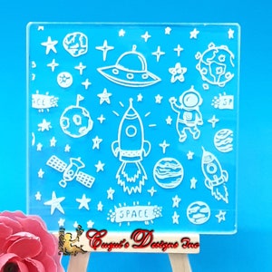 May include: Clear silicone stamp with a space theme. The stamp features a rocket, astronaut, UFO, planets, stars, and the word "SPACE".