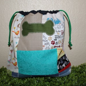 Puede incluir: Una bolsa de cordón de tela patchwork con una aplicación de hueso verde y la palabra "Find" bordada en ella. La bolsa está hecha de varias telas coloridas con diferentes patrones, incluyendo pájaros, huellas de patas y huesos.