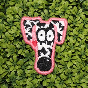 May include: A black and white zebra-patterned iron-on patch with pink stitching. The patch features a cartoon zebra with large eyes and a pink nose.