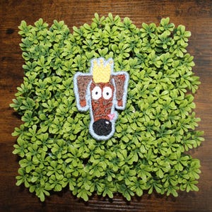 May include: Embroidered patch featuring a cartoon dog with a crown. The dog has brown fur, large white eyes, and a black nose. The patch is outlined in blue and set against a bed of green foliage on a wooden surface.
