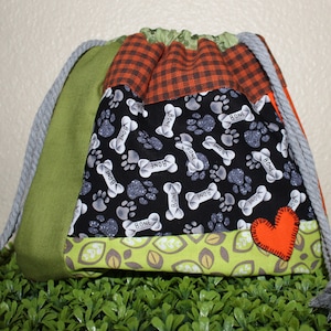Pode incluir: Uma bolsa com cordão com um design de patchwork. A bolsa apresenta um tecido preto com pegadas brancas e gráficos de ossos, tecido xadrez laranja e marrom e tecido verde. Um pequeno coração laranja é costurado na frente. O cordão é cinza.