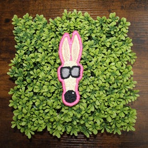 May include: A whimsical patch featuring a cartoon rabbit head with sunglasses. The rabbit has pink ears and a pink outline, with a black nose and sunglasses. The patch is set against a backdrop of small green leaves on a wooden surface.