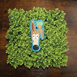 May include: Embroidered patch featuring a cartoon dog with large eyes, a brown ear, and a black nose. The dog is holding a carrot with green leaves. The patch has a blue outline and a small pink heart. The patch is set on a square of green foliage.