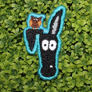 Op de afbeelding: Zwart-witte geborduurde patch van een hondenkop met grote ogen en een kleine bruine uil die er bovenop zit. De patch is omrand met turquoise.