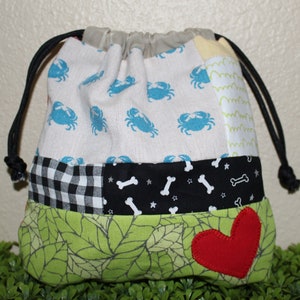 Pode incluir: Uma bolsa de cordão de retalhos com um padrão de folhas verdes, tecido xadrez preto e branco, um tecido preto com desenhos de ossos e estrelas brancas e um tecido branco com desenhos de caranguejos azuis. A bolsa tem um coração vermelho costurado na frente.