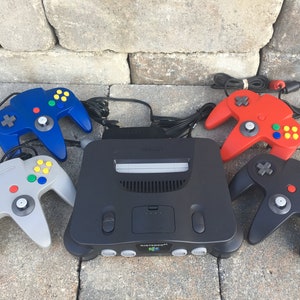 May include: A black Nintendo 64 console with four controllers. The controllers are gray, blue, red, and black. The console has a gray cartridge slot and a silver Nintendo logo.