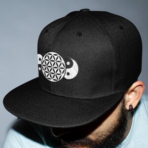 May include: A black and white patterned baseball cap with a flower of life and yin yang symbol. The cap is worn by a person with a beard.